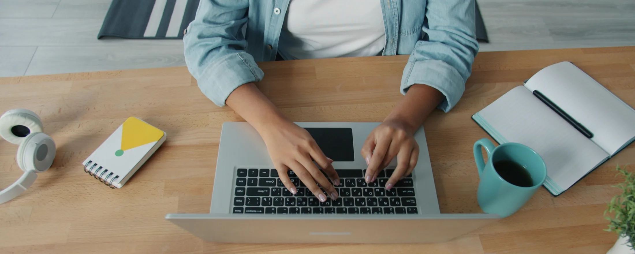 A person typing on a laptop at a wooden desk, seen from above, with a notebook and a mug of coffee